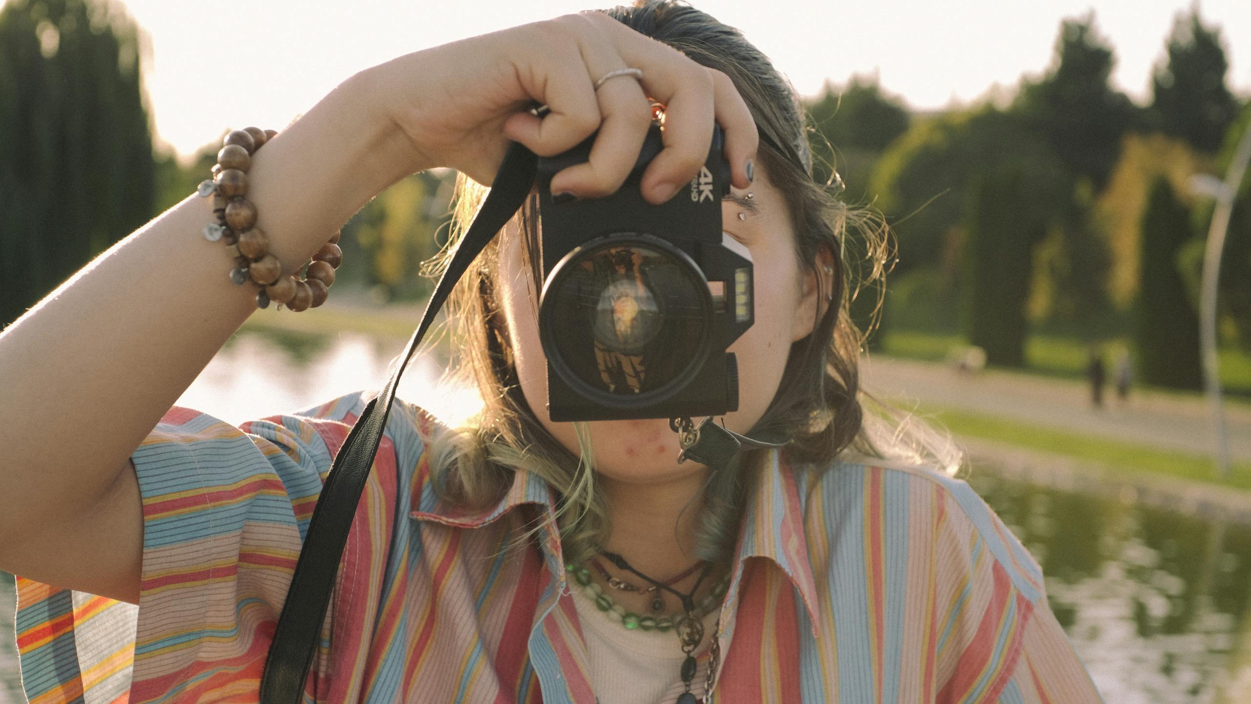 A person in a striped shirt uses a camera outdoors near water and trees.